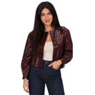A woman modeling a cropped, glossy dark burgundy faux leather jacket over a cream tank top and dark blue jeans, set against a plain white background.