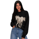 A woman wearing a black knit sweater with a large beige and gray horse graphic on the front, paired with blue jeans, against a plain white background.