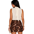 A woman shown from the back wearing a sleeveless cream blouse with a large bow at the neckline and a brown sequined mini skirt, holding a metallic clutch against a plain white background.