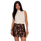 A woman modeling a sleeveless cream mock-neck top and a brown sequined mini skirt, holding a small beige clutch against a plain white background.
