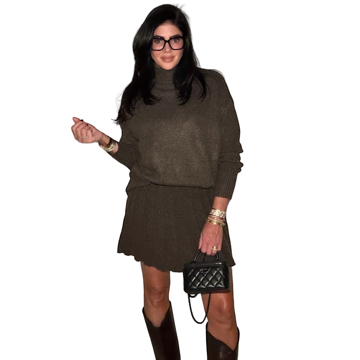 Woman wearing a brown sweater and skirt set with a black handbag against a plain wall.