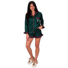 A woman modeling a dark green satin pajama set with red and black plaid trim, featuring a long-sleeve button-front top and matching shorts, paired with beige slippers on a white background.