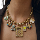 A close-up of a model wearing a gold charm necklace with multiple decorative pendants, including religious icons, a teddy bear, a heart, and an envelope charm.