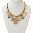 A gold-tone charm necklace displayed on a cream-colored mannequin bust. The necklace features a chunky link chain and multiple dangling charms, including two gold-tone Miraculous Medal pendants, a teddy bear charm, a small yellow heart, a light green square charm, and a white envelope charm.