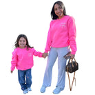 Woman and child wearing matching pink sweatshirts with a logo, standing next to a handbag on a white background.