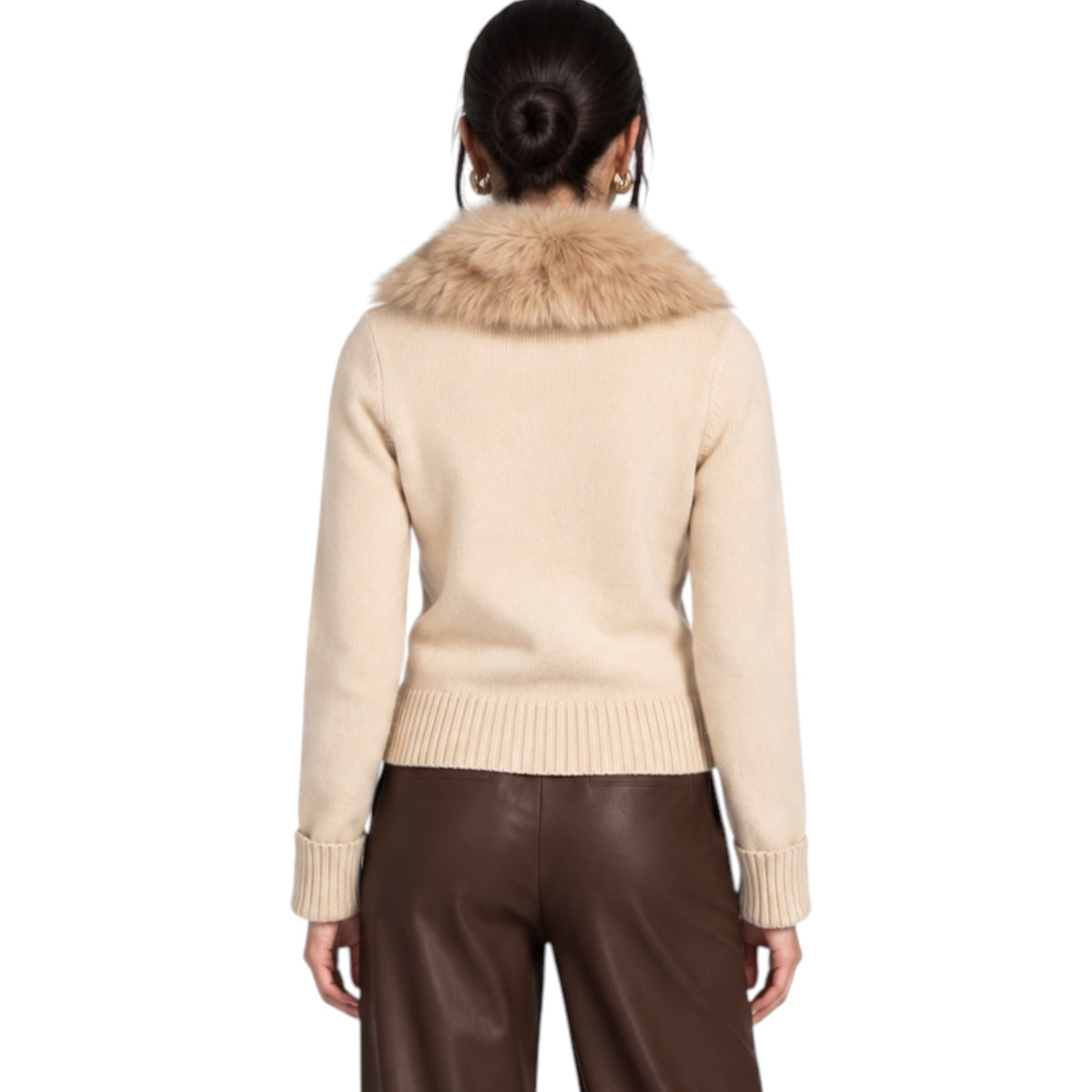 A woman viewed from the back wearing a beige knit sweater with a large tan faux-fur collar and dark brown pants, against a plain white studio background.