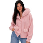 A woman wearing a pink fuzzy knit cardigan with a large faux-fur trimmed hood, drawstrings, and pom-pom details, paired with blue jeans against a plain white background.