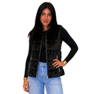 A woman modeling a black faux fur puffer vest with a front zipper over a black long-sleeve top and light blue jeans, set against a plain white background.
