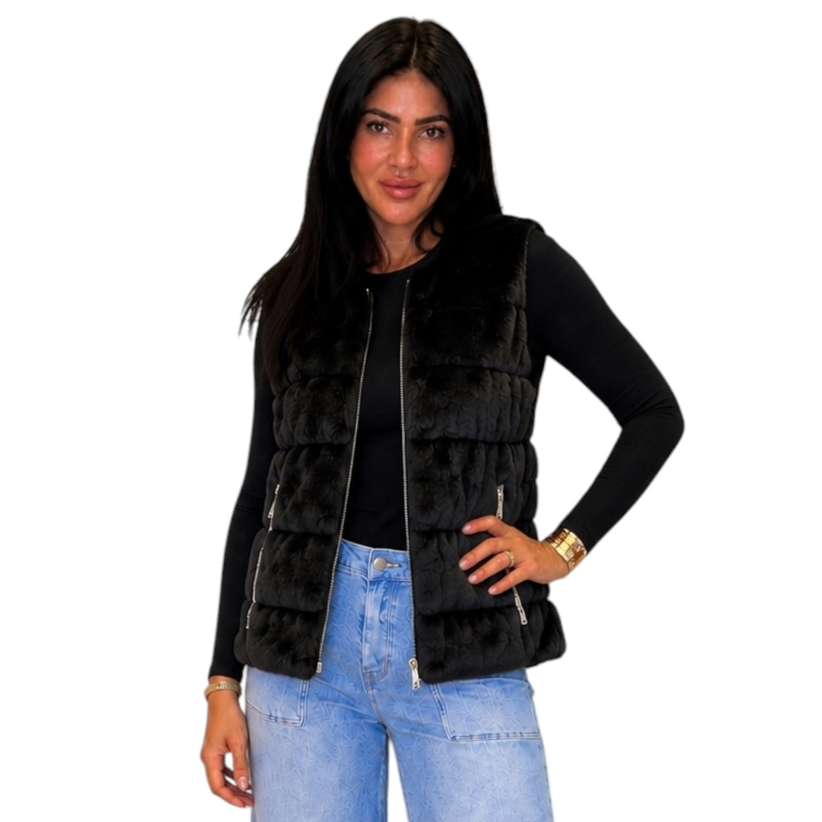 A woman modeling a black faux fur puffer vest with a front zipper over a black long-sleeve top and light blue jeans, set against a plain white background.