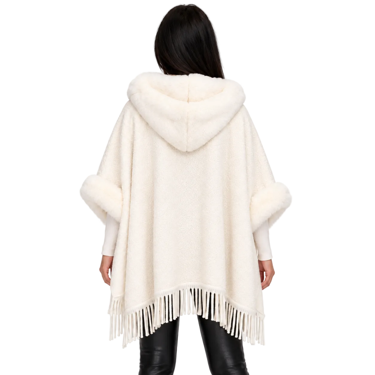 A woman viewed from behind wearing a cream-colored hooded poncho with faux fur trim on the hood and sleeves, and fringe along the hem, paired with black pants against a plain white background.