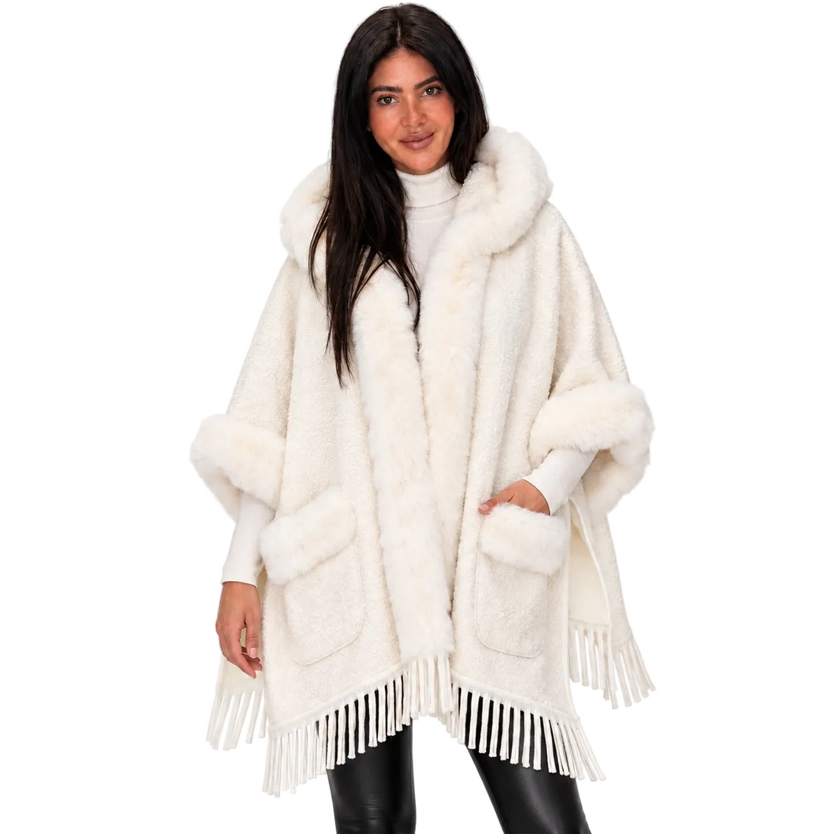 A woman modeling a fluffy off-white faux fur poncho with a large hood, front pockets, and fringe trim, styled over a white turtleneck and black pants against a plain white background.