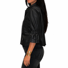 A woman shown in side profile wearing a black faux-leather cropped jacket with a gathered elastic waist and elbow-length sleeves, paired with dark jeans and gold bracelets, against a plain white background.
