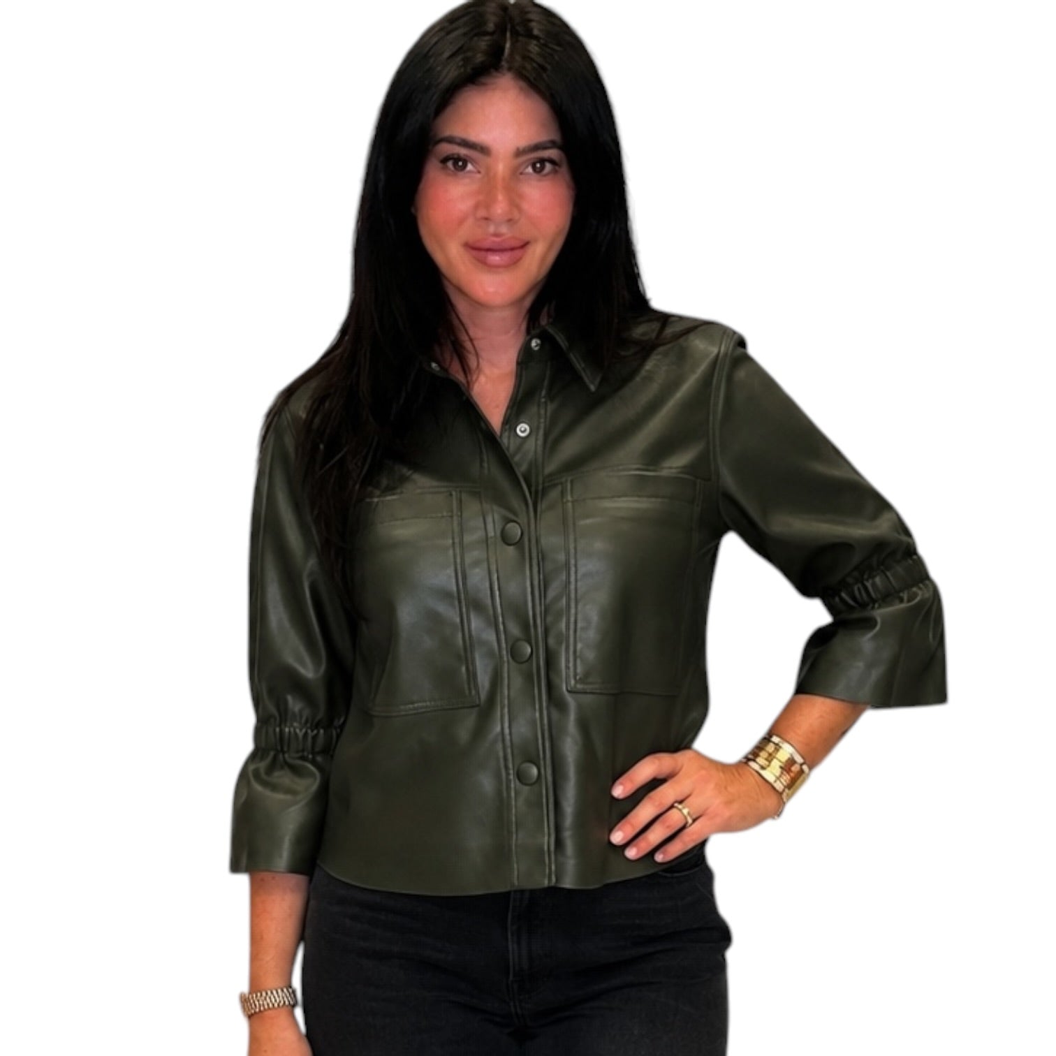 A woman modeling a dark olive green faux leather button-up shirt jacket with chest pockets and gathered cuffs, paired with black pants, against a plain white background.