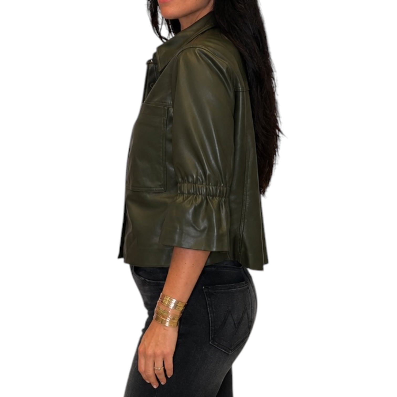 A woman shown in side profile wearing an olive green faux-leather jacket with a gathered waist and three-quarter sleeves, paired with black jeans against a plain white background.