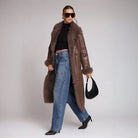 Woman wearing a long brown fur-trimmed coat, black top, blue jeans, and black shoes on a plain background.