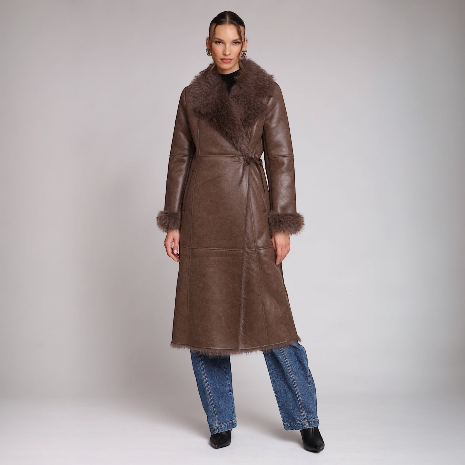 Woman wearing a long brown leather coat with fur trim on a plain background