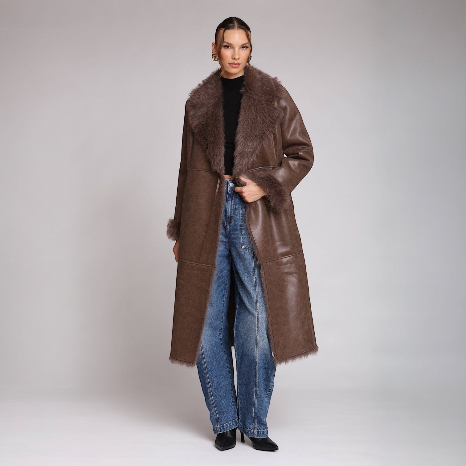 Person wearing a long brown coat with fur trim on a plain background
