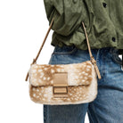 A person wearing blue jeans and an olive green shirt carries a small shoulder bag with a brown and white spotted faux-fur texture, tan leather trim, and a tan buckle closure.