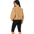 A woman shown from the back wearing a tan cropped jacket with a high collar and black capri pants, photographed against a plain white background.
