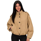 A woman modeling a cropped tan utility jacket with a high stand collar, dark buttons, and shoulder epaulets, paired with black pants against a plain white background.