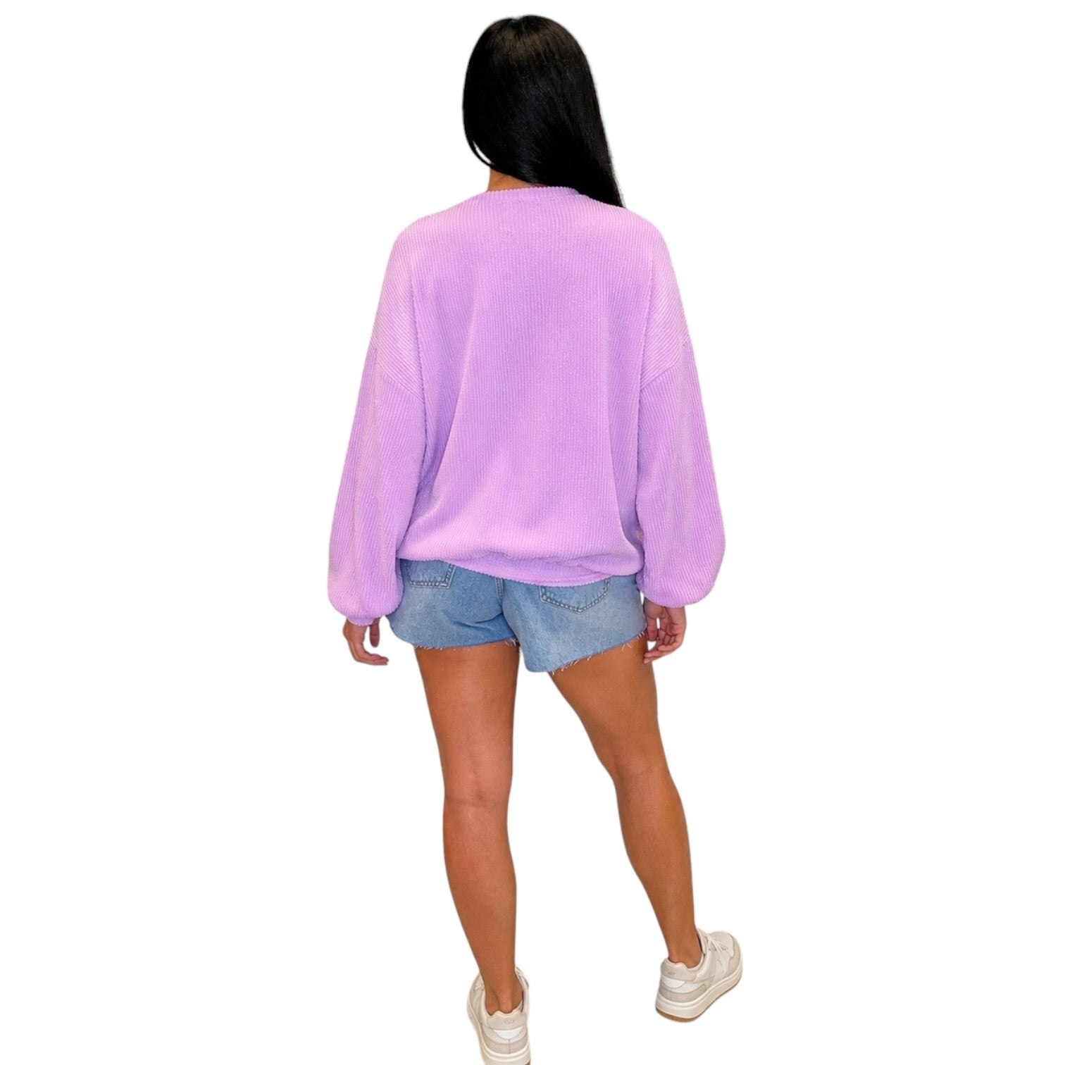 A woman shown from the back wearing a lavender-purple long-sleeve sweatshirt, denim shorts, and white sneakers against a plain white background.