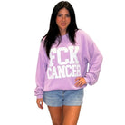 A woman modeling a lavender purple ribbed knit sweatshirt with 'FCK CANCER' printed in bold white block letters across the front, paired with denim shorts against a plain white background.