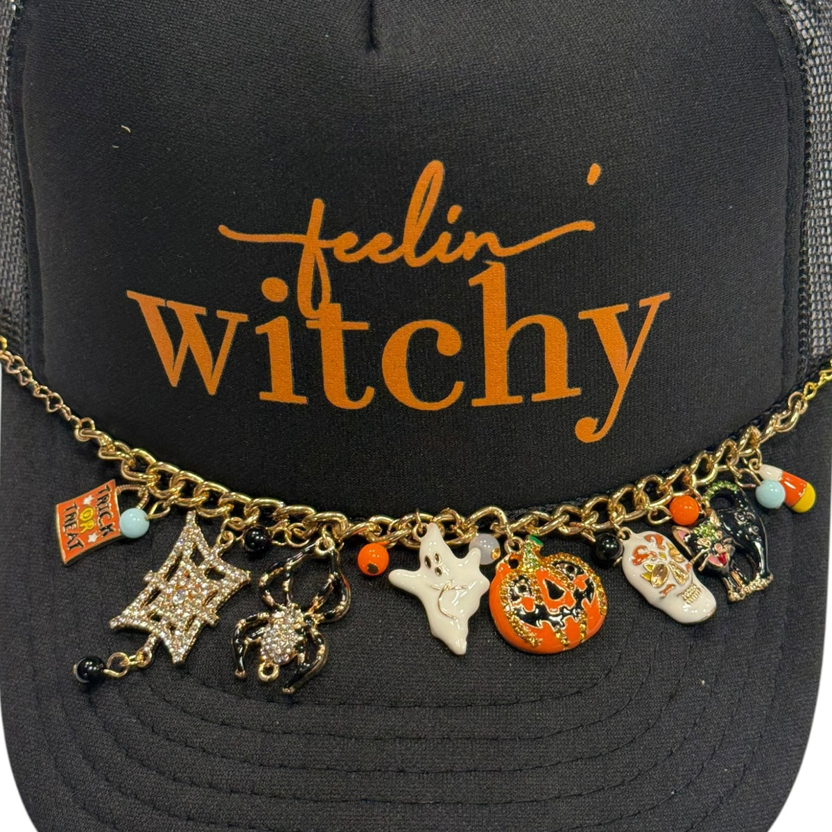 A black trucker hat with orange embroidered text reading 'feelin' witchy' and a gold-tone charm necklace draped across the brim. The charms include a spider, ghost, pumpkin, skull, candy corn, and small beads.
