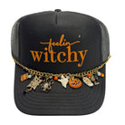 A black trucker hat with orange embroidered text reading 'feelin' witchy' and a gold charm chain draped across the brim, featuring Halloween-themed charms including a spider, witch hat, pumpkin, and boot.