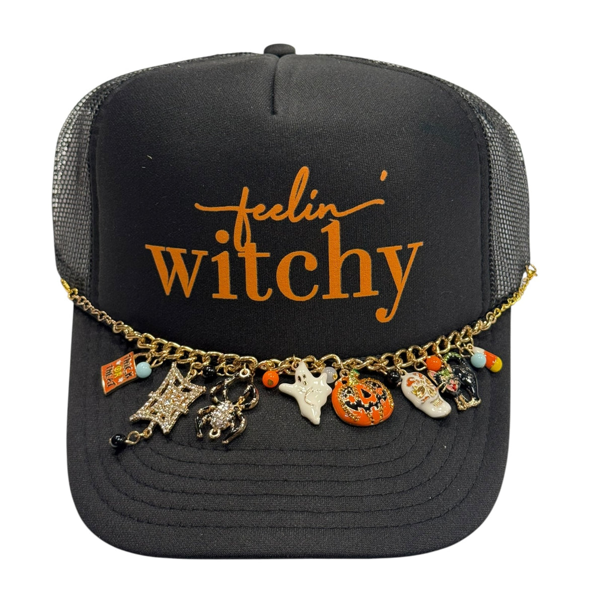 A black trucker hat with orange embroidered text reading 'feelin' witchy' and a gold charm chain draped across the brim, featuring Halloween-themed charms including a spider, witch hat, pumpkin, and boot.