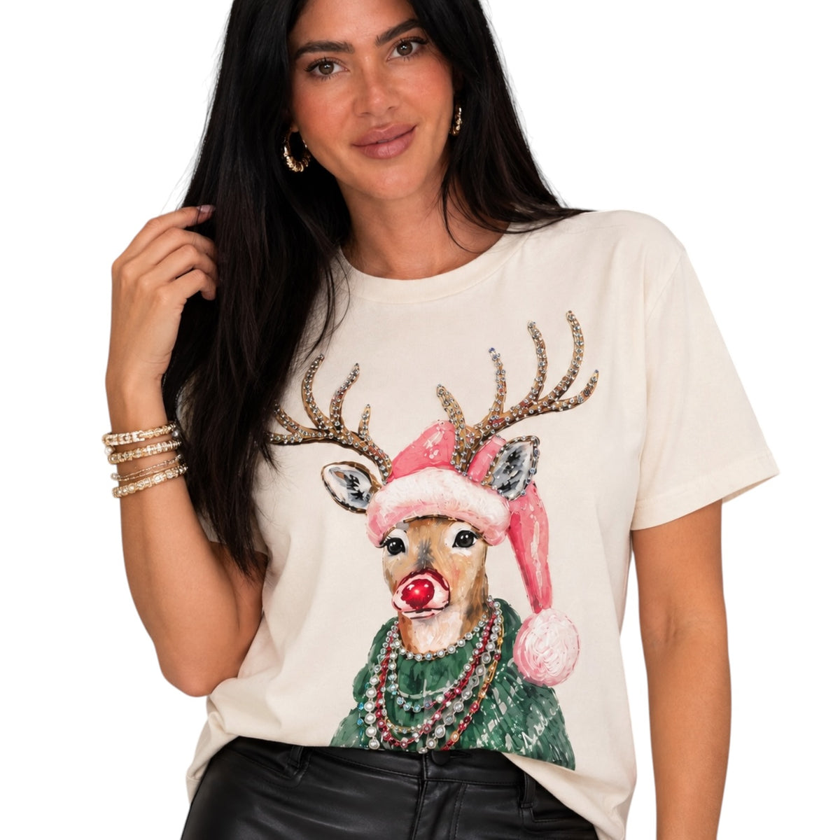 A woman wearing a cream-colored t-shirt with a festive graphic of a reindeer wearing a pink Santa hat, green sweater, and red nose. The background is solid white.