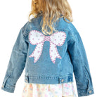 Child wearing a denim jacket with a large pink bow on the back, standing against a light pink background.