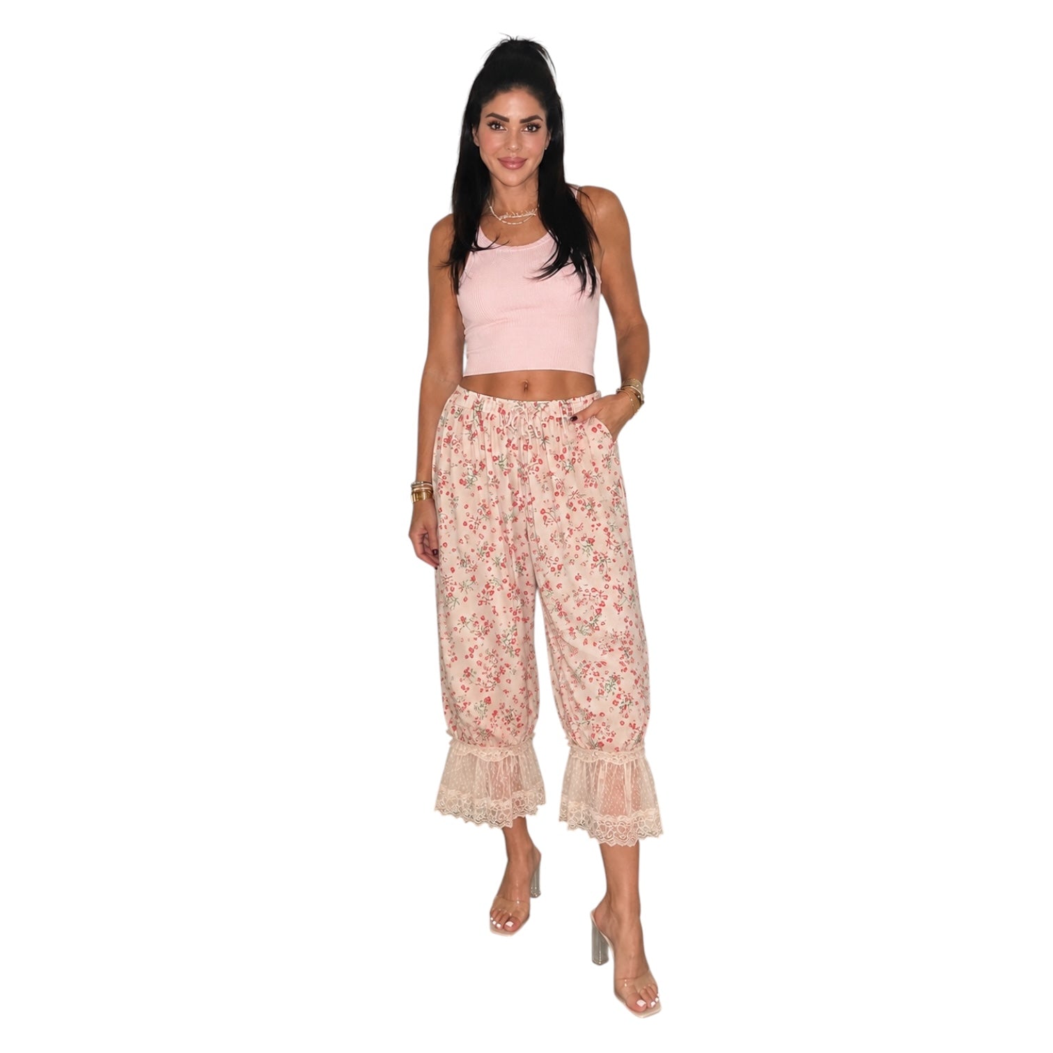 A woman modeling a two-piece outfit consisting of a light pink sleeveless crop top and high-waisted, wide-leg floral pants with lace ruffle hems, set against a plain white background.