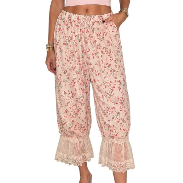 A woman modeling a two-piece outfit consisting of a light pink sleeveless crop top and high-waisted, wide-leg floral pants with lace ruffle hems, set against a plain white background.