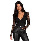 A woman modeling a black long-sleeve top with sheer lace sleeves and a scalloped V-neckline, paired with black faux-leather pants, against a plain white background.