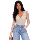 A woman modeling a beige lace long-sleeve bodysuit with a V-neckline, paired with light blue jeans, against a plain white background.