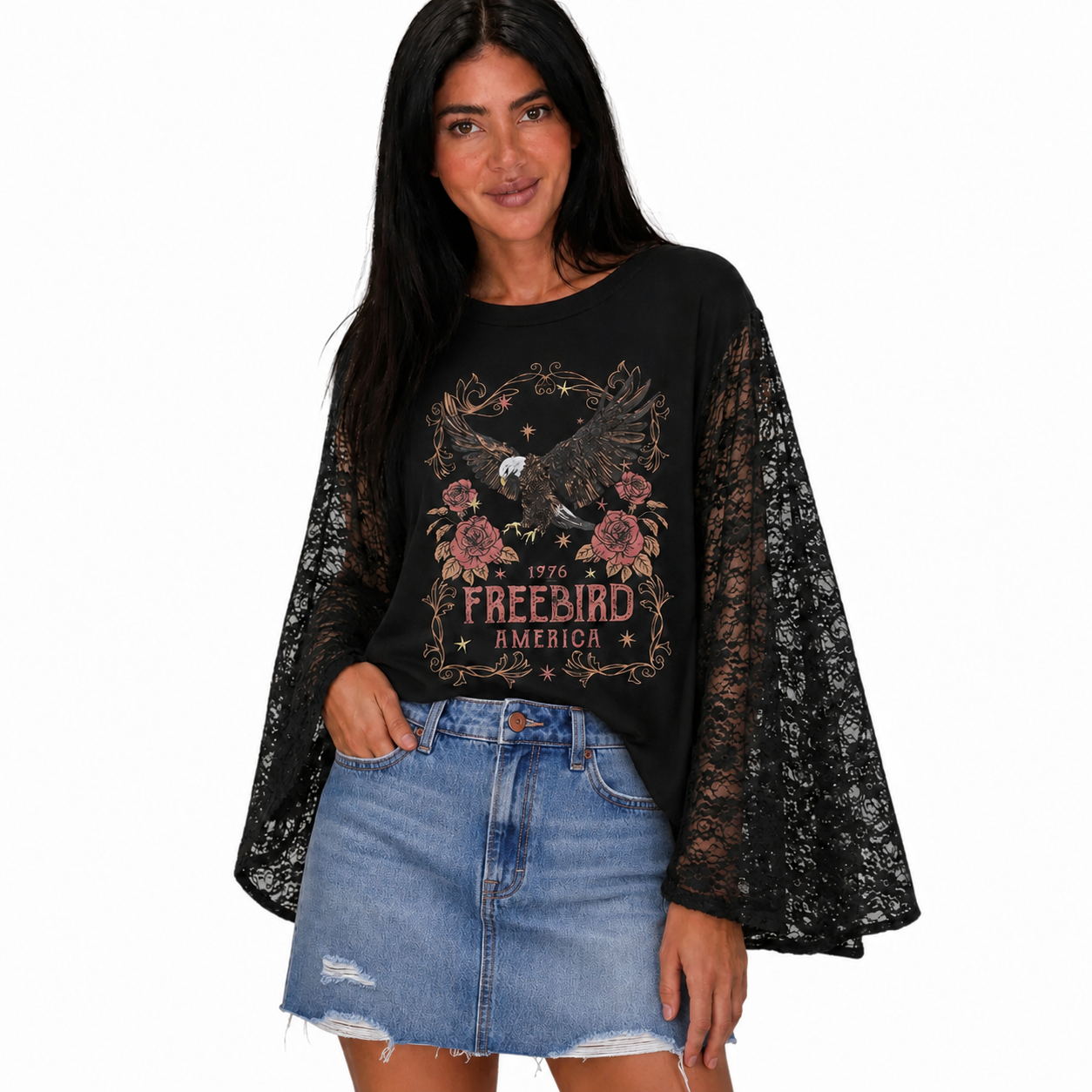 A woman modeling a black long-sleeve top with sheer lace bell sleeves and a graphic print on the front featuring an eagle, flowers, and the text '1976 FREEBIRD AMERICA', paired with a light blue denim skirt against a plain white background.