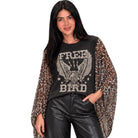 A woman modeling a black graphic t-shirt with a distressed eagle design and the words 'FREE BIRD', layered under a sheer leopard-print long-sleeve top, paired with black faux-leather pants, against a plain white background.