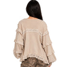 A woman viewed from the back wearing a beige long-sleeve top with layered ruffles and lace trim, paired with camouflage pants, against a plain white background.