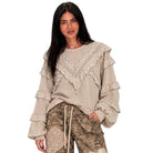 A woman modeling a beige long-sleeve top with layered ruffles and lace trim, paired with camouflage pants, against a plain white background.