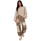 A woman modeling a bohemian-style outfit featuring a beige ruffled long-sleeve top and brown camouflage patchwork cargo pants with crochet details, paired with beige sneakers, against a solid white background.