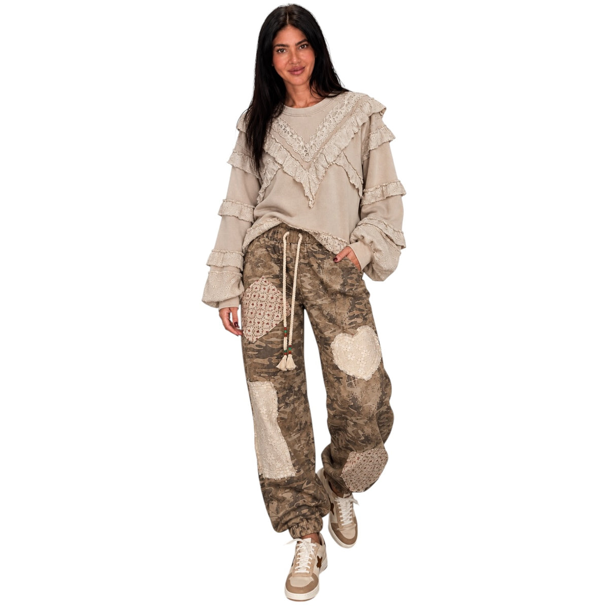 A woman modeling a bohemian-style outfit featuring a beige ruffled long-sleeve top and brown camouflage patchwork cargo pants with crochet details, paired with beige sneakers, against a solid white background.
