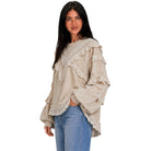 A woman modeling a beige long-sleeve top with layered ruffles and lace trim, paired with blue jeans, against a plain white background.