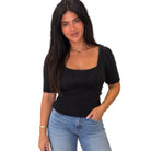 A woman modeling a black short-sleeve knit top with a square neckline and ribbed hem, paired with light blue jeans, against a plain white background.