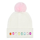 An off-white ribbed knit pom-pom beanie featuring a fluffy light pink pom-pom on top and a row of colorful faux gemstones on the folded cuff.