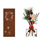 Holiday-themed reed diffuser set with a clear glass bottle, black reeds, green pine branches, red berries, a gingerbread man ornament, and a red-and-green ribbon, shown beside its brown decorative box featuring a wreath design.