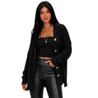 A woman modeling a black open-front knit cardigan with gold-tone buttons, worn over a black tube top and black faux-leather pants, against a plain white background.