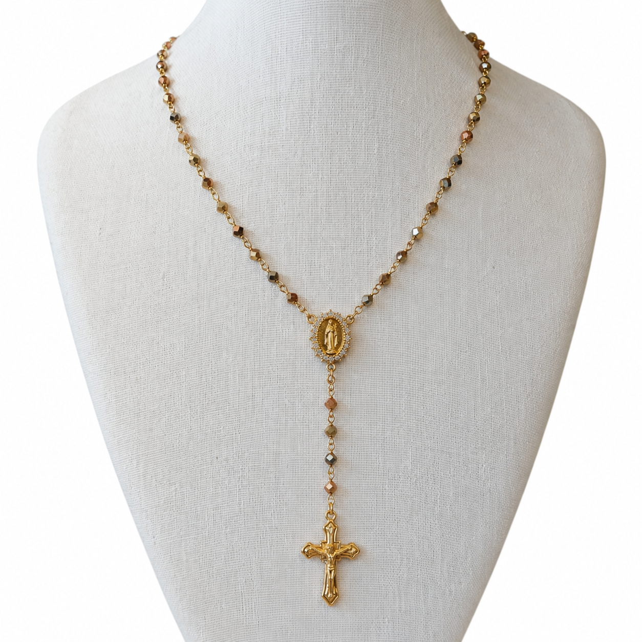 A gold-toned rosary necklace displayed on a white fabric mannequin bust against a white background. The necklace features small round beads, a central oval medallion, and a dangling crucifix pendant.
