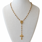 A gold-toned rosary necklace displayed on a white fabric mannequin bust against a white background. The necklace features small round beads, a central oval medallion, and a dangling crucifix pendant.