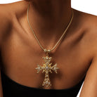 A gold cross pendant necklace adorned with clear crystals, worn by a model on a white background.