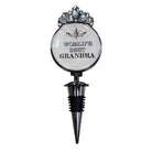 A novelty wine bottle stopper designed as a round medallion with the text 'WORLD'S BEST GRANDMA' in black lettering on a white background, topped with a silver rhinestone crown, and fitted with a black and silver threaded stopper base.